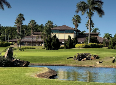 Casarão de telhado marrom entre palmeiras, com gramado e um pequeno lago